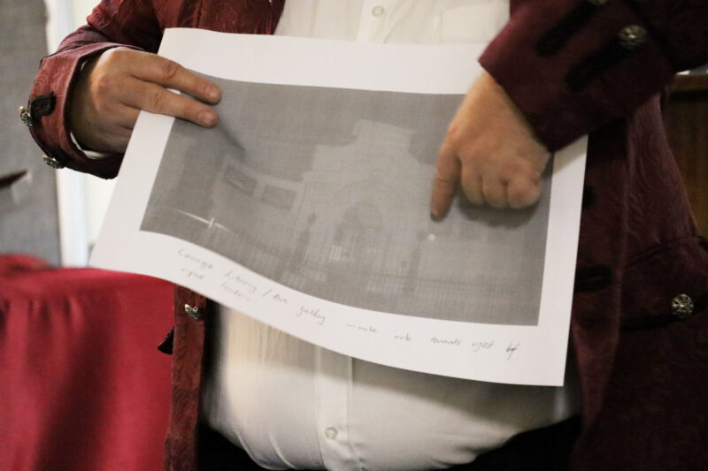 A grey photo showing a foggy building. The man is pointing at a round light with his left index finger. The photo has a white boarder around it.