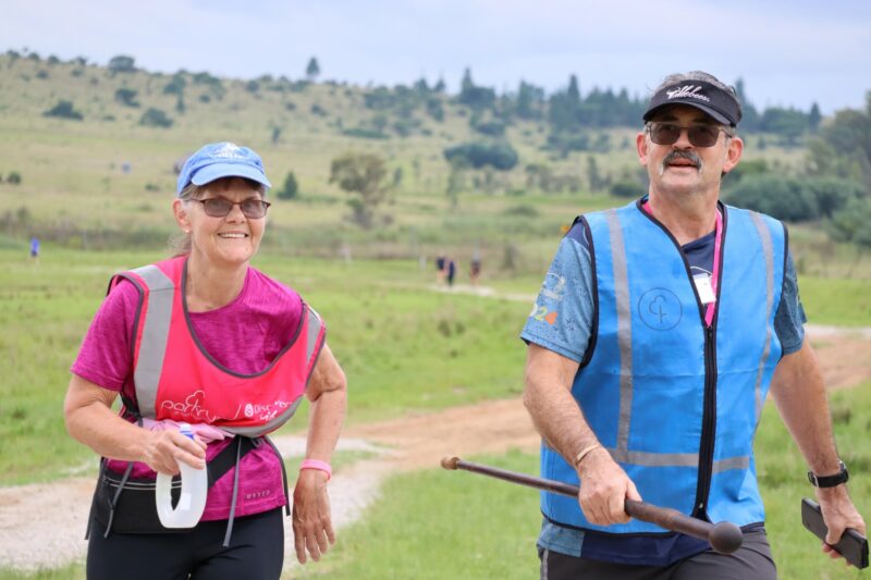 A man and a woman is walking down a dirt road. THe woman is wearing a pink reflective vest; the man is wearing a blue reflective vest. The man has a stick in his right hand. The woman is carrying a water bottle in her right hand. THere are mountains in the background.