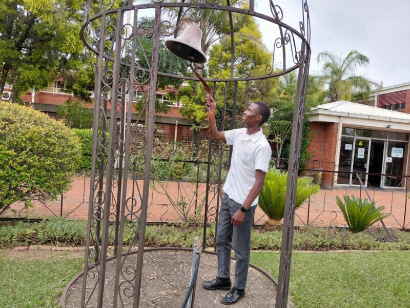 A photo showing learners from the Bridging Group 2025 taking turns to ring a ceremonial bell outdoors at Tugela Pre-Vocational School. Each photo features one or a small group of learners standing beneath the metal bell structure in a garden area with trees and school buildings in the background.