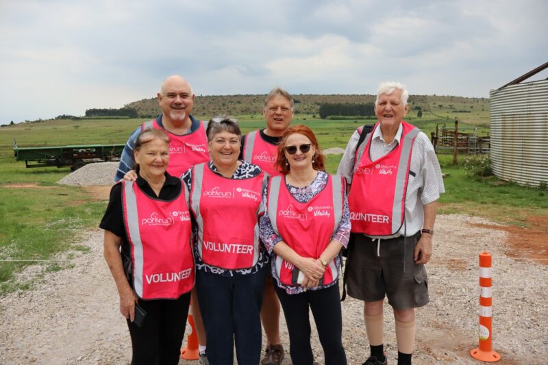 Six people are standing in a group. THey are wearing pink reflective vests. They are outside, and the background is filled with a farmy/mountainous background. The sky is cloudy.