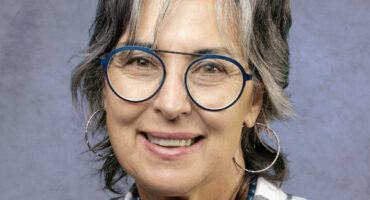 An elderly teacher is dressed in grey and blue clothing poses in front of a grey background.