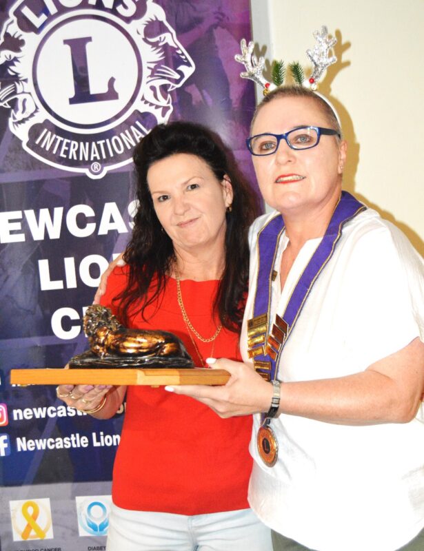 Two women posing in front of a Lions international Banner holding a trophy.