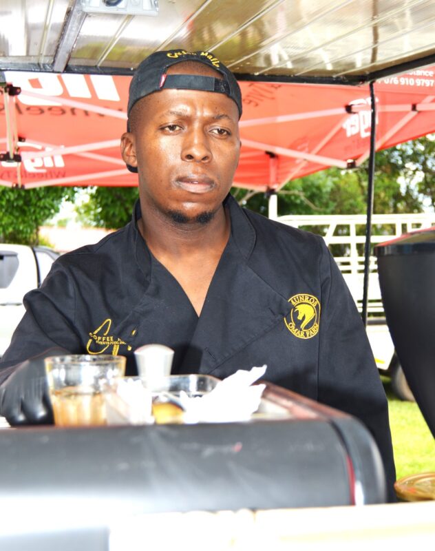 One man is photographed serving beverages during a market day under a red gazebo.