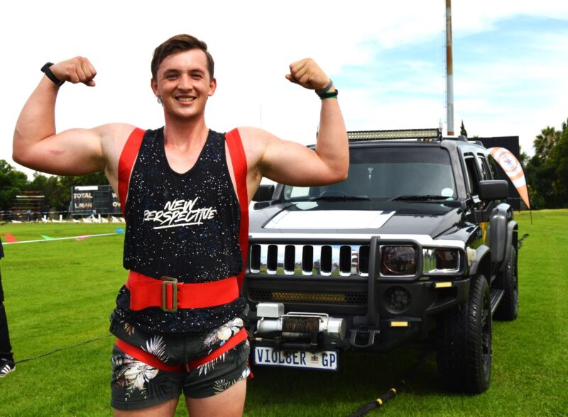 A young man dressed in black and red, flexing his muscles while getting ready to pull a black SUV.