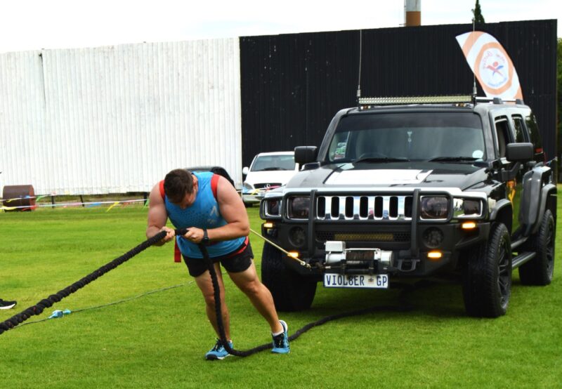 A young man wearing blue and red, has a rope around his waist while he is pulling a black SUV vehicle during a strong man competition.