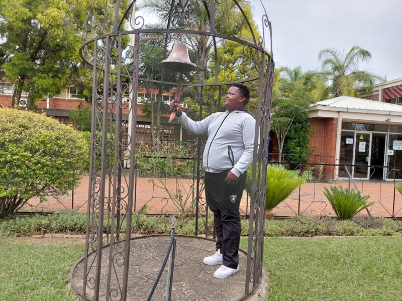 A photo showing learners from the Bridging Group 2025 taking turns to ring a ceremonial bell outdoors at Tugela Pre-Vocational School. Each photo features one or a small group of learners standing beneath the metal bell structure in a garden area with trees and school buildings in the background.