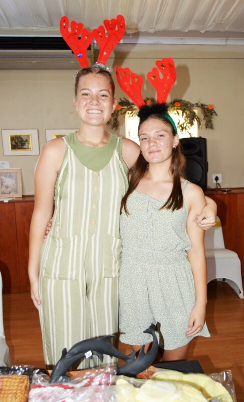 A photo of two young girls posing at a market stand wearing reindeer ears.