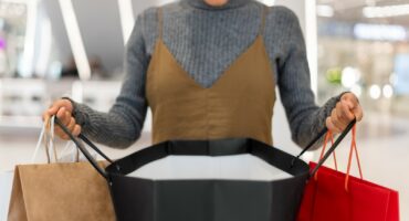 A women is photographed holding three shopping bags coloured red, brown and black. She appears to be in a retail store.