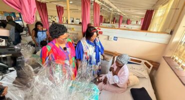 Two women are seen standing by a new mother and her baby, bearing a gift in a public hospital setting.