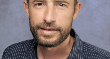 A man is looking at the camera. He has a beard and short, brown hair. He is wearing a buttoned-up shirt. The background is grey-blue.