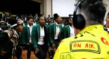 A radio presenter with a yellow, red, white, and black shirt has headphones on his head. He is looking at school children in front of him. The children are wearing white and green. They are standing in front of a school hall with a white screen in the bachround.