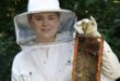 A lady in a bee protection suit is holding a honey store. Her head protection has net in front of it. She is holding the honey store in a vertical position. Bees, wax and honey is visable on the honey store. Her bee protection suit is white. She is wearing gloves. There are green leaves and trees in the background.