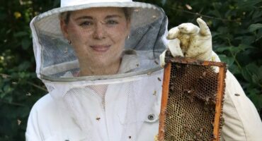 A lady in a bee protection suit is holding a honey store. Her head protection has net in front of it. She is holding the honey store in a vertical position. Bees, wax and honey is visable on the honey store. Her bee protection suit is white. She is wearing gloves. There are green leaves and trees in the background.