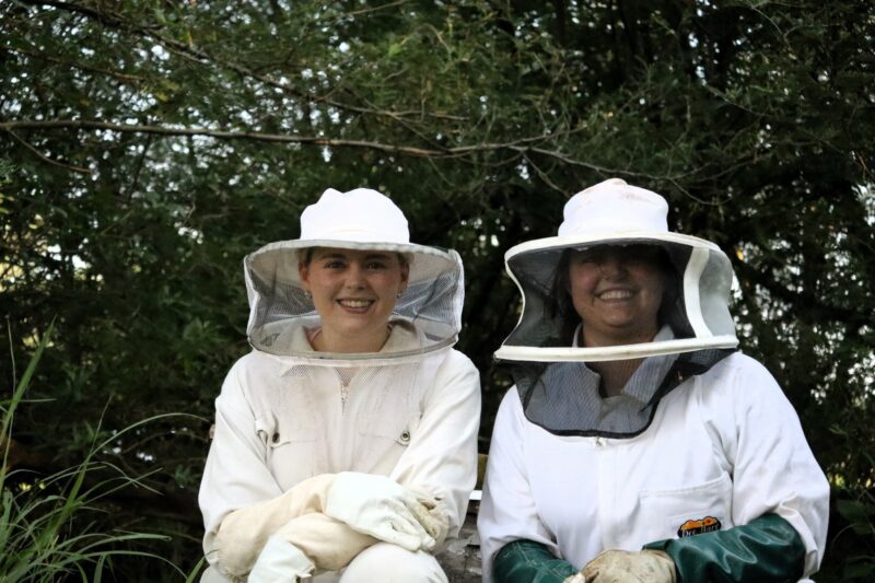 Two people in white bee suits are smiling at the camera. They are kneeling next to each other. The one lady is wearing green gloves, the other one is wearing white gloves. There are a lot of trees behind them. The trees are green.