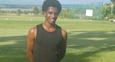 Image of an African man dressed in black vest and black pants smiling and posing on green grass.