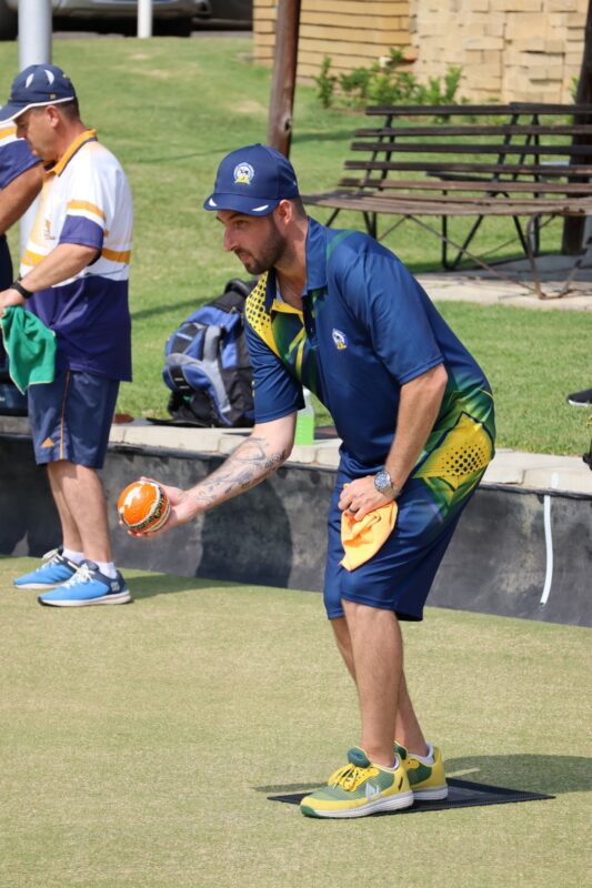 A man wearing a bowls outfit, with a cap on his head. He is holding a bowling ball in his right hand. He is wearing his team's uniform that is green, blue, and yellow. He is standing on a green bowling pitch.