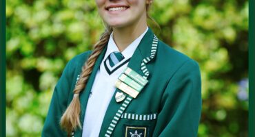 A school portrait of a high school student. The student is wearing a green blazer, white jersey, and green and white tie. There are multiple badges pinned to her blazer's collar. She is smiling, and her hair is braided to the one side of her shoulder. There are green leaves in a blurred background.