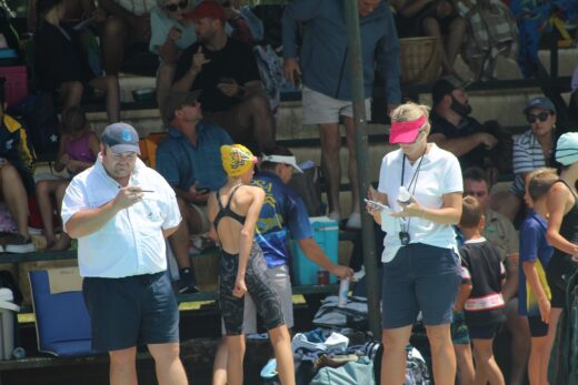 A man and a woman dressed in white shirts and blue short pants standing next to the pool while checking results.