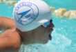 Image of a swimmer with a white swim cap with blue writing and goggles, taking a breath of air before continuing with breast stroke.