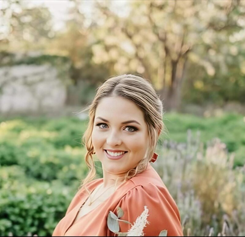 A portrait photo of a young teacher taken outside in a garden setting.