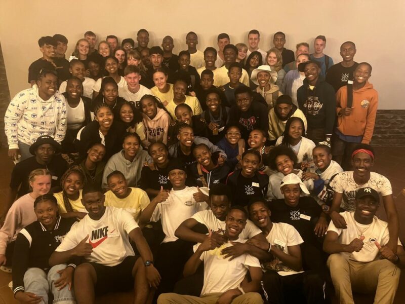 A big group of the Representative Council of Learners (RCL) are smiling and looking at the camera. The photo has a yellowish tint. There is a light-coloured wall in the blackground. The stdents are wearing different coloured shirts.