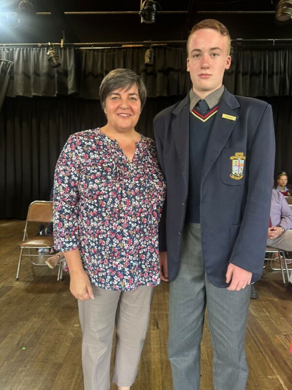 A tall high school student standing next to a shorter teacher. The student is wearing a blazer, jersey, long pants and tie. He has a badge pinned to his jacket. The teacher is wearing a printed blouse.