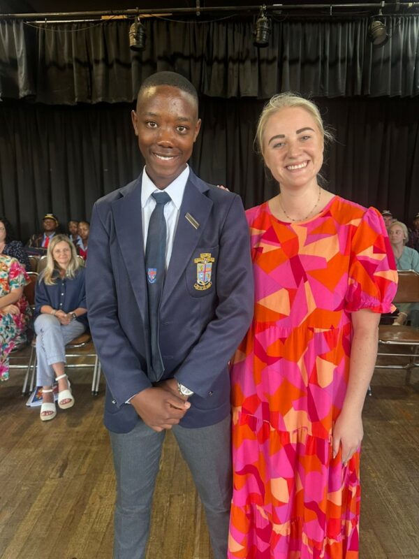 A high school student is standing next to a teacher. The student is a bit taller than the teacher. The student is wearing a blazer and tie of the same colour. The teacher is wearing a pink, red and orange coloured dress. Both are smiling. There are people sitting in the background.