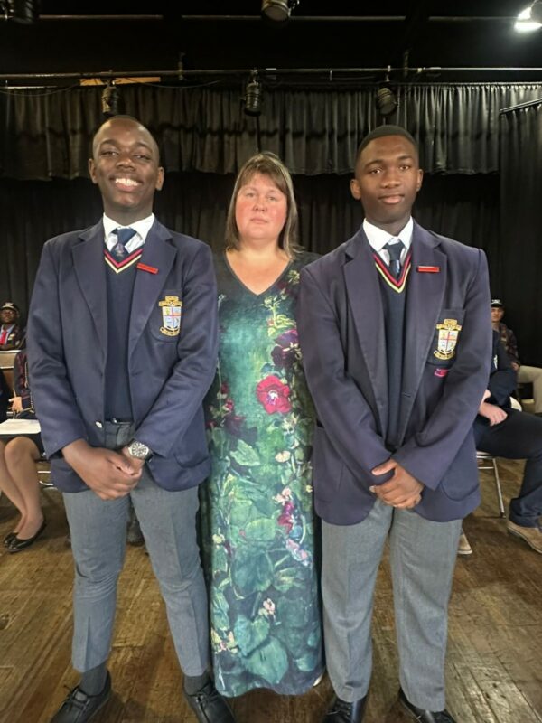 Two high school students are standing on either side of the teacher. Both are taller than the teacher. The students are wearing a blazer, jersey, and tie of the same bolour. Both students' hands are folded in front of them. Each has a red badge binned to the blazer's collar. The teacher is wearing a green-ish floral dress.