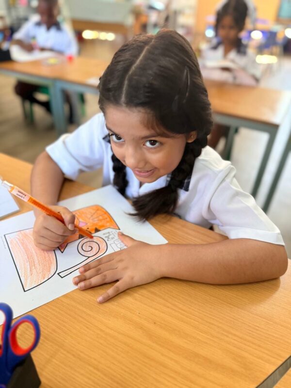 A photo of a grade 1 learner posing at her desk while she colours in a picture.