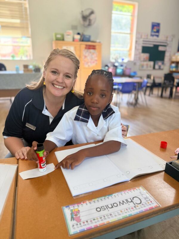A Grade 1 learner poses for a photo with her teacher. In the picture is a colour-in book and crayons on her desk.