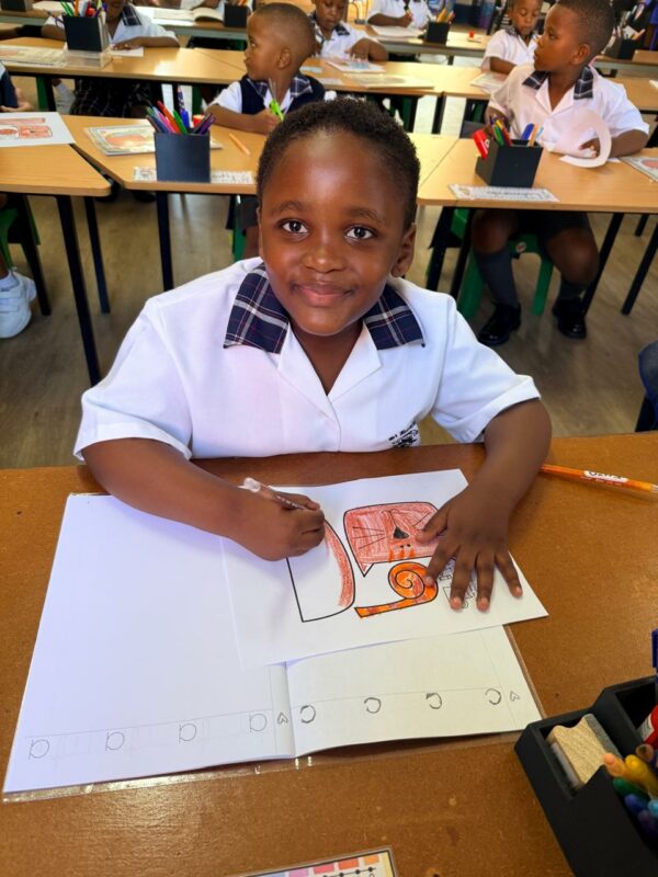 A photo of a grade 1 learner posing at her desk while she colours in a picture.