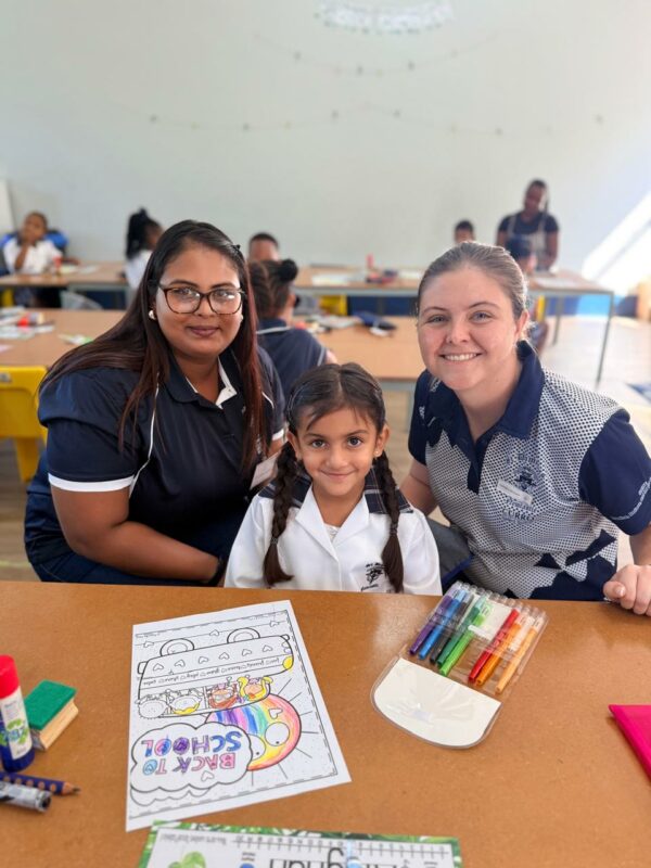 A Grade 1 learner poses for a photo with two of her teachers. In the picture is a colour-in book and crayons on her desk.