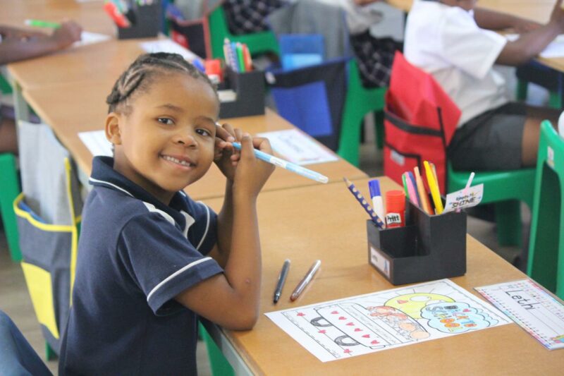 A photo of a grade 1 learner posing at her desk while she colours in a picture.