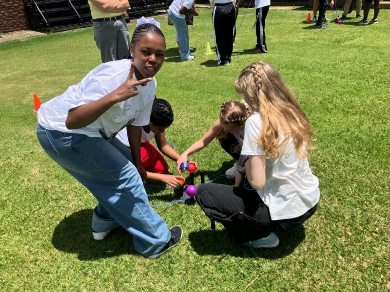 High school students are outdoors on green grass. They are kneeling in front of something that is read, reaching it with their hands. A student is looking at the camera and showing a "peace" sign with the back of her hand toward the camera. It is sunny.