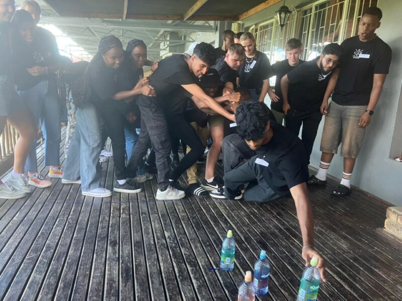 High school learners with black t-shirts on. They are under a roof on a wooden surface. One student is placing a bottle on the floot next to other bottles which seems to be filled with water. The students are wearing jeans and sneakers.