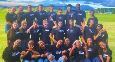 The Elected Committee group. Some students are standing, kneeling and sitting. It is day-time with clouds in the sky. There are mountains visable in the background. The students are wearing black shirts with name tags and white printing on the right side.