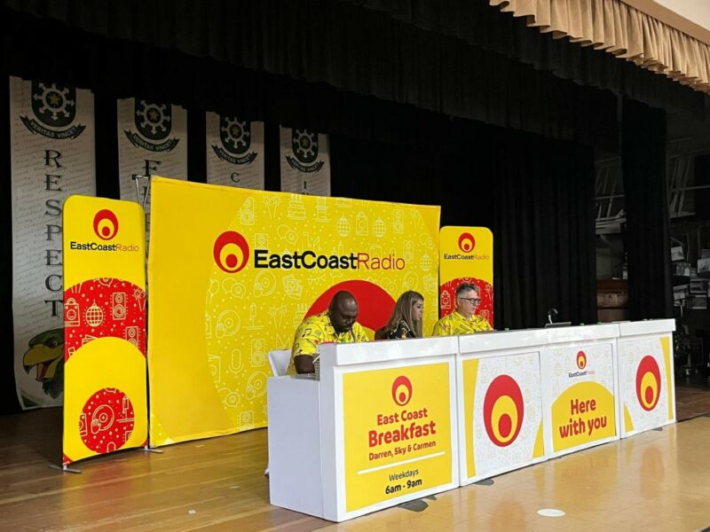 A radio station booth on a school stage. The radio presenters are sitting in a row in front of a red, yellow and black banner with the radio station's name and logo on it. They are sitting behind a white, yellow and red commentary box with the radio station's name and logo on it. All three presenters are wearing headphones.