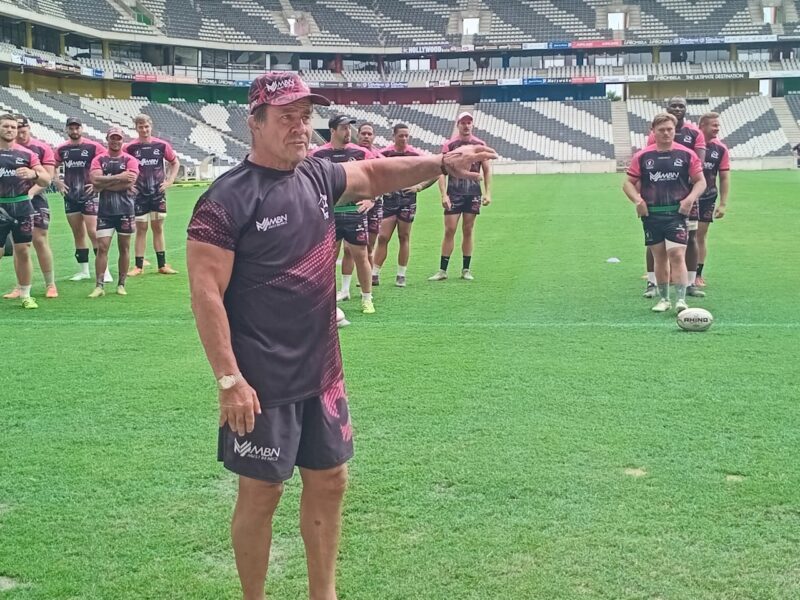 A whole group of rugby players standing on a rugby field with the coach pointing forward.
