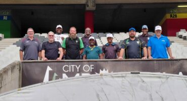 A row of male rugby coaches pose for a photo in front of the Pumas Rugby Stadium.
