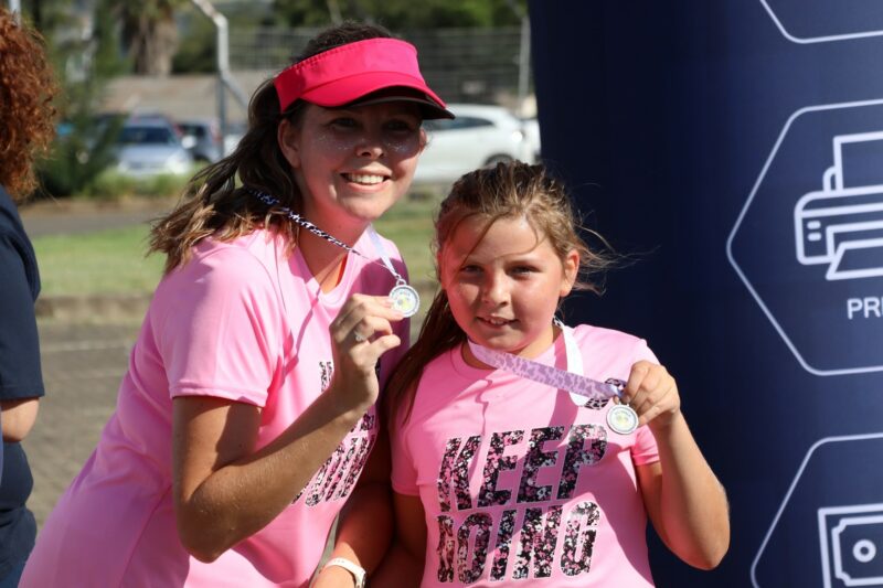 Two ladies embracing the Valentine's Day spirit with their pink t-shirts. They are holding medals after completing the Lions Club 4 kilometer walk for cancer. The older lady has a hat on her head and the sun is shining.