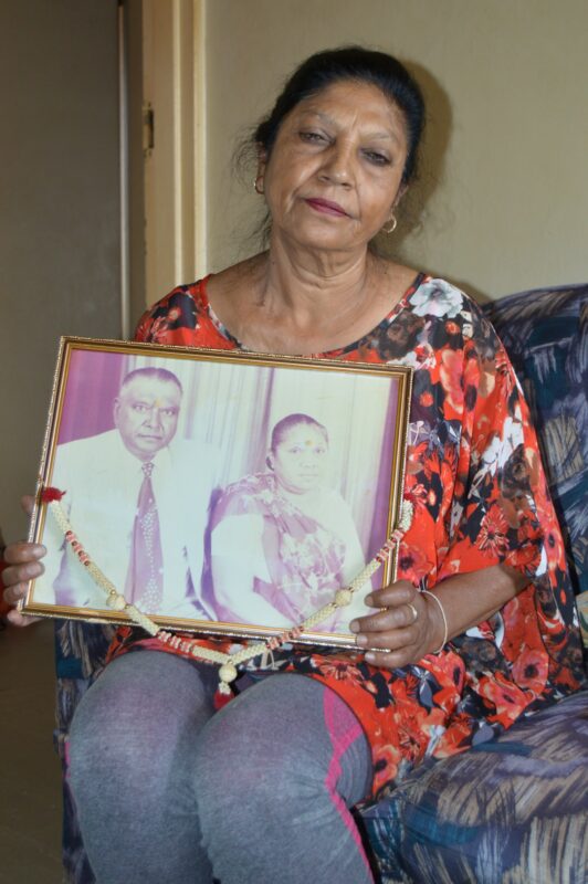 An elderly woman is photographed sitting in a chair holding a photo of her parents.