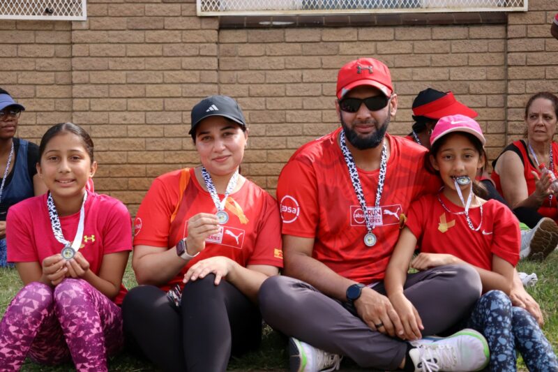 The Docrat Family is embracing the Valentine's Day spirit by wearing pink and read. The are posing for a photo after completing the 4 kilometer walk for cancer. Mother, father and one daughter is wearing caps on their heads. The father has a pair of sunglasses on his nose. They are sitting in front of a facebrick wall.