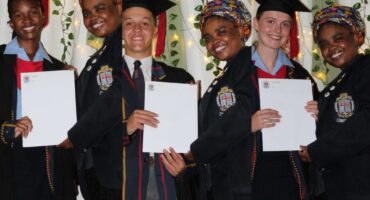 A collage of three students and a teacher holding academic certificates. All three students have graduation hats on their heads and they are wearing school blazers. The teacher is wearing a school blazer too, with a headscarf on her head. There are fairy lights and greenery in the background.