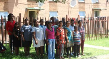 A group of tenants of the Suryaville Municipal Flats pose outside the building under a tree.