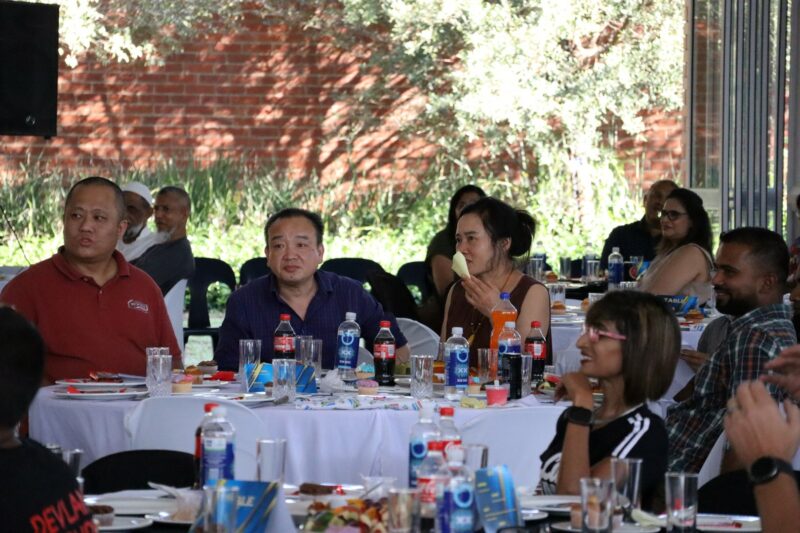 Asian people sitting at a table with food and drink on the table. They are sitting inside a school hall with trees in the background. The Asian people are looking in front of them. The tablecloth on the table is white. It is day-time.
