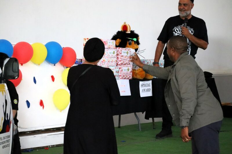 A man is throwing darts at multi-coloured balloons. There are two people watching the man throw the dart. The balloons are attched to a white board. There is a white wall in the background.