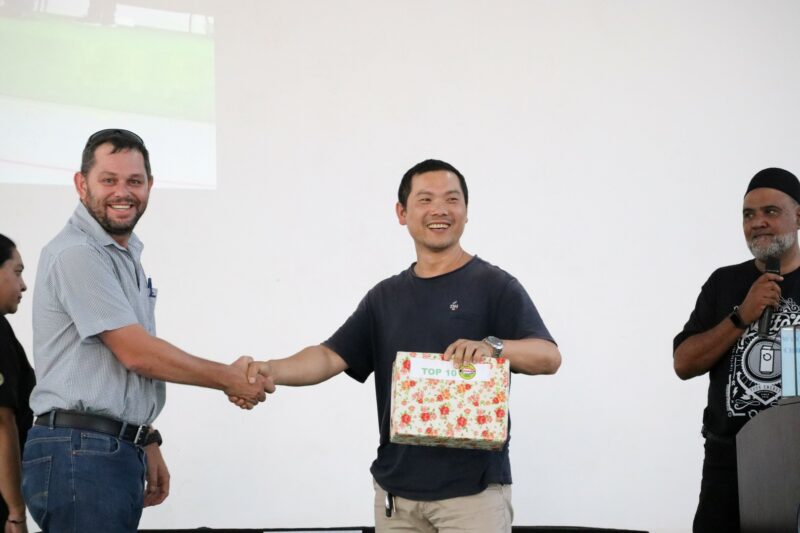 A white man is shaking an Asian man's hand. The Asian man is holding a box in his left hand. There is an Indian man next to him, speaking into a microphone. There is a screen projected against the white wall in the background.