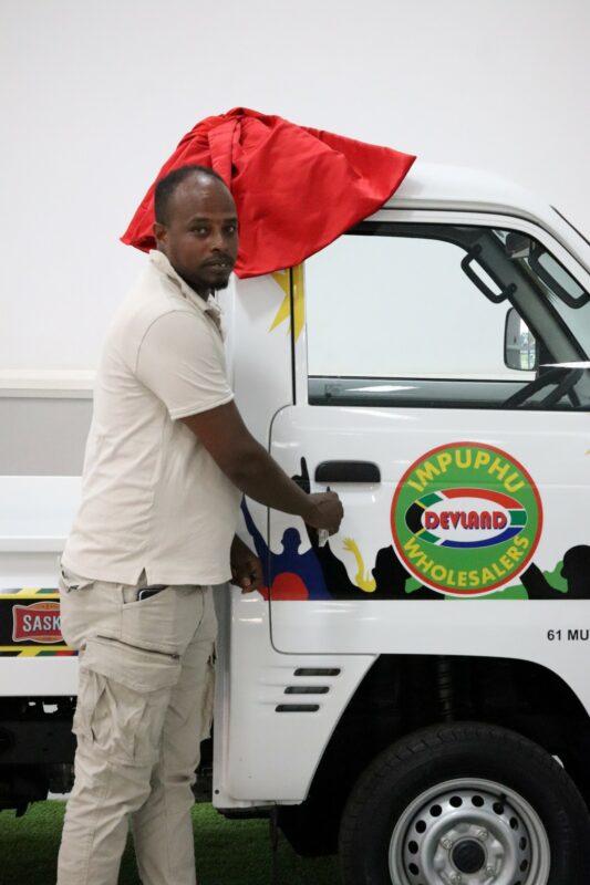 A man is standing in front of a white vehicle. The vehilce has many stickers on it. There is a red ribbon attached to the vehicle. There is a white wall behind the vehicle. The man is dark-skinned and wearing light colours.