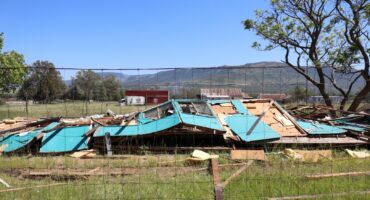 A collapsed classroom at Utrecht Primary School. The classroom is now behind a fence to secure further damages and or loss of property. The classroom was a turqoise colour. It is a very bright day, with mountains in the bachground. The sky is a deep blue.