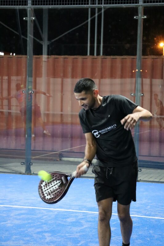 A young man is playing paddle. He is facing the camera and hitting the ball. The racquet is in his right hand and his left arm is in the air. He is wearing all-black with white writing on his shirt. The court surface is blue and there is a brown container behind him. He is playing at night.